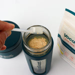 VocalGuard Glass Bottle — leveling a scoop of powder over the open bottle, VocalGuard pouch nearby.