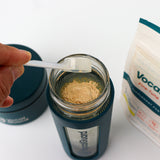 VocalGuard Glass Bottle — leveling a scoop of powder over the open bottle, VocalGuard pouch nearby.