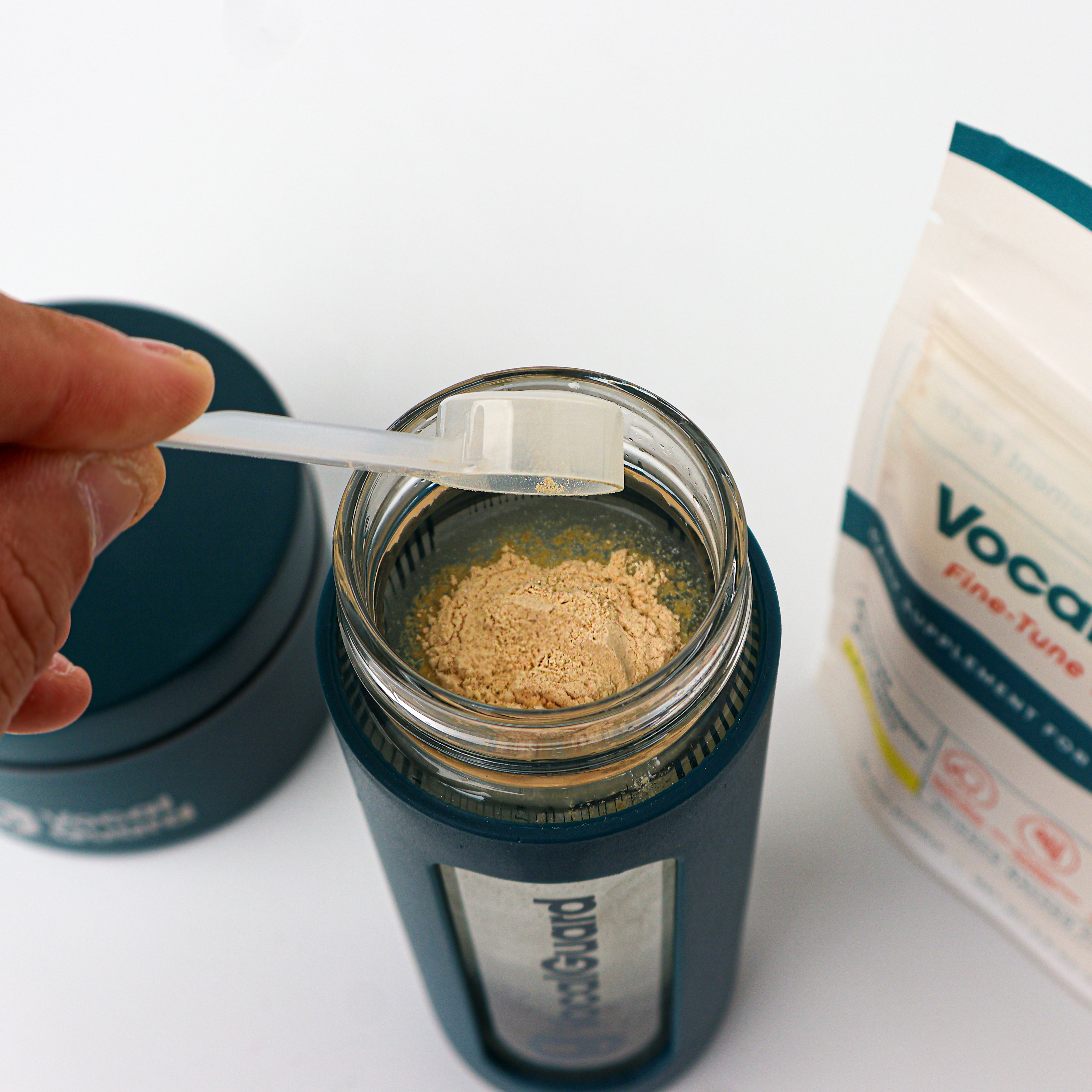 VocalGuard Glass Bottle — leveling a scoop of powder over the open bottle, VocalGuard pouch nearby.
